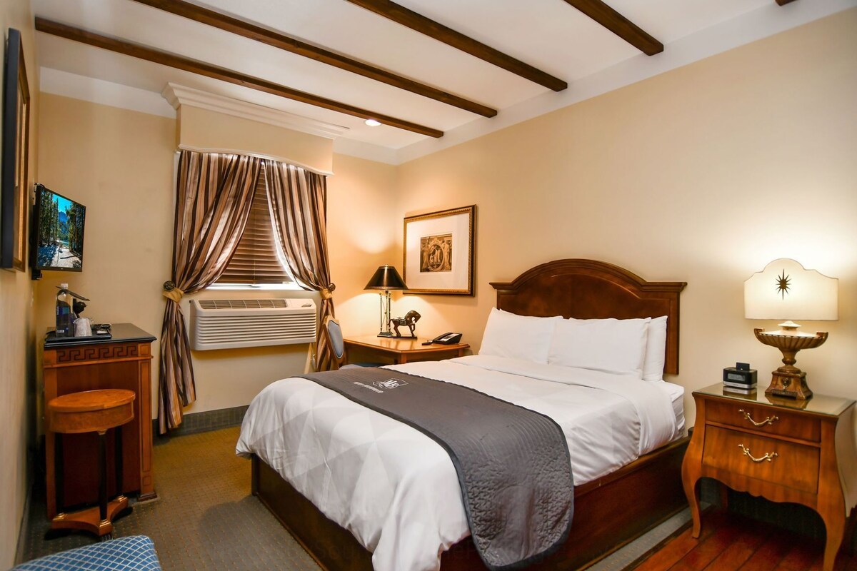 Habitación de hotel con cama, mesitas de noche, escritorio, aire acondicionado, ventana con cortinas y un cuadro en la pared.