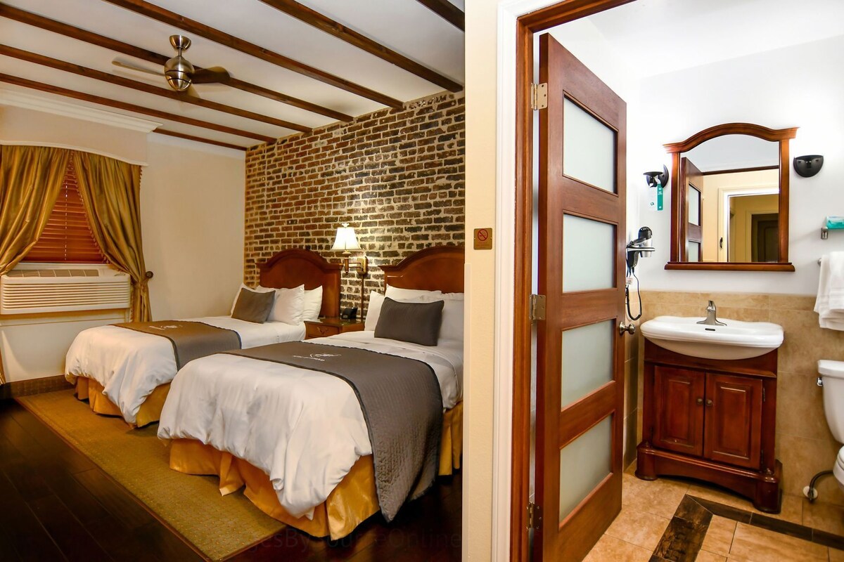 Habitación de hotel con dos camas, pared de ladrillo, techo de madera, baño con lavamanos y espejo, y una puerta.