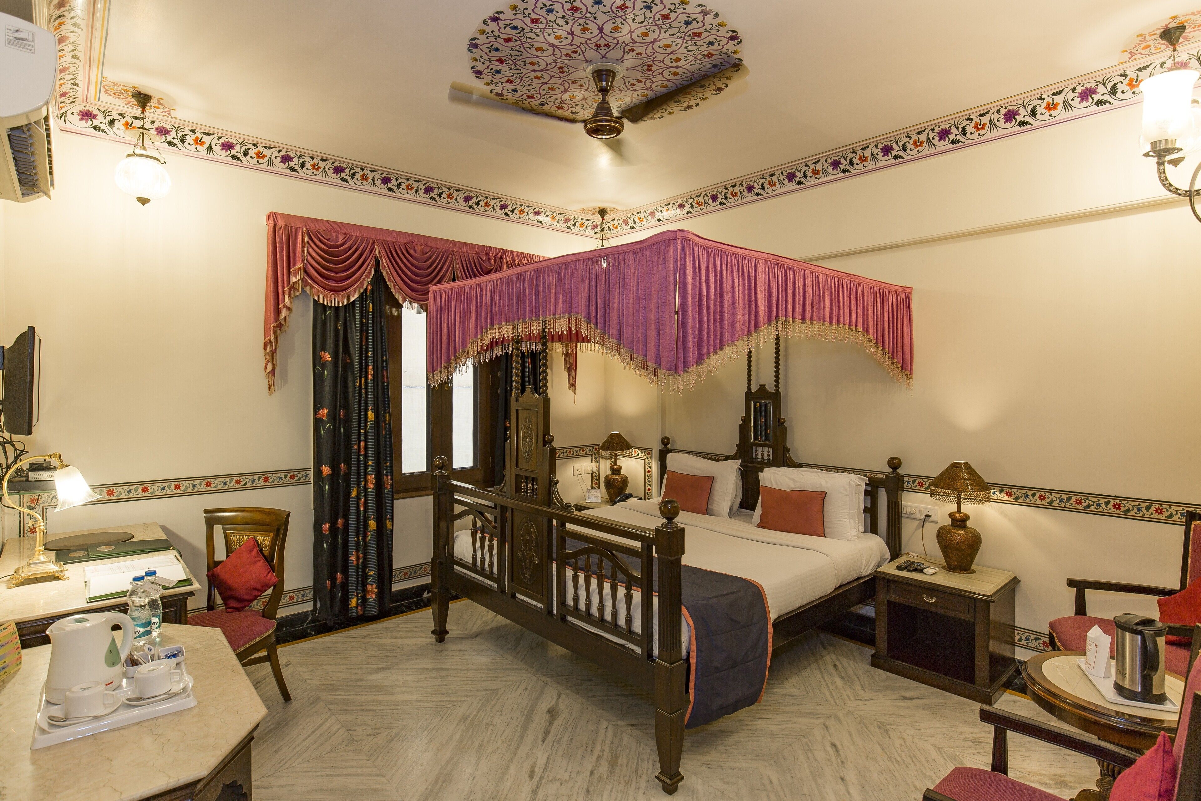 A four-poster bed with a canopy, a desk with a lamp, a chair, and a small table with a kettle.
