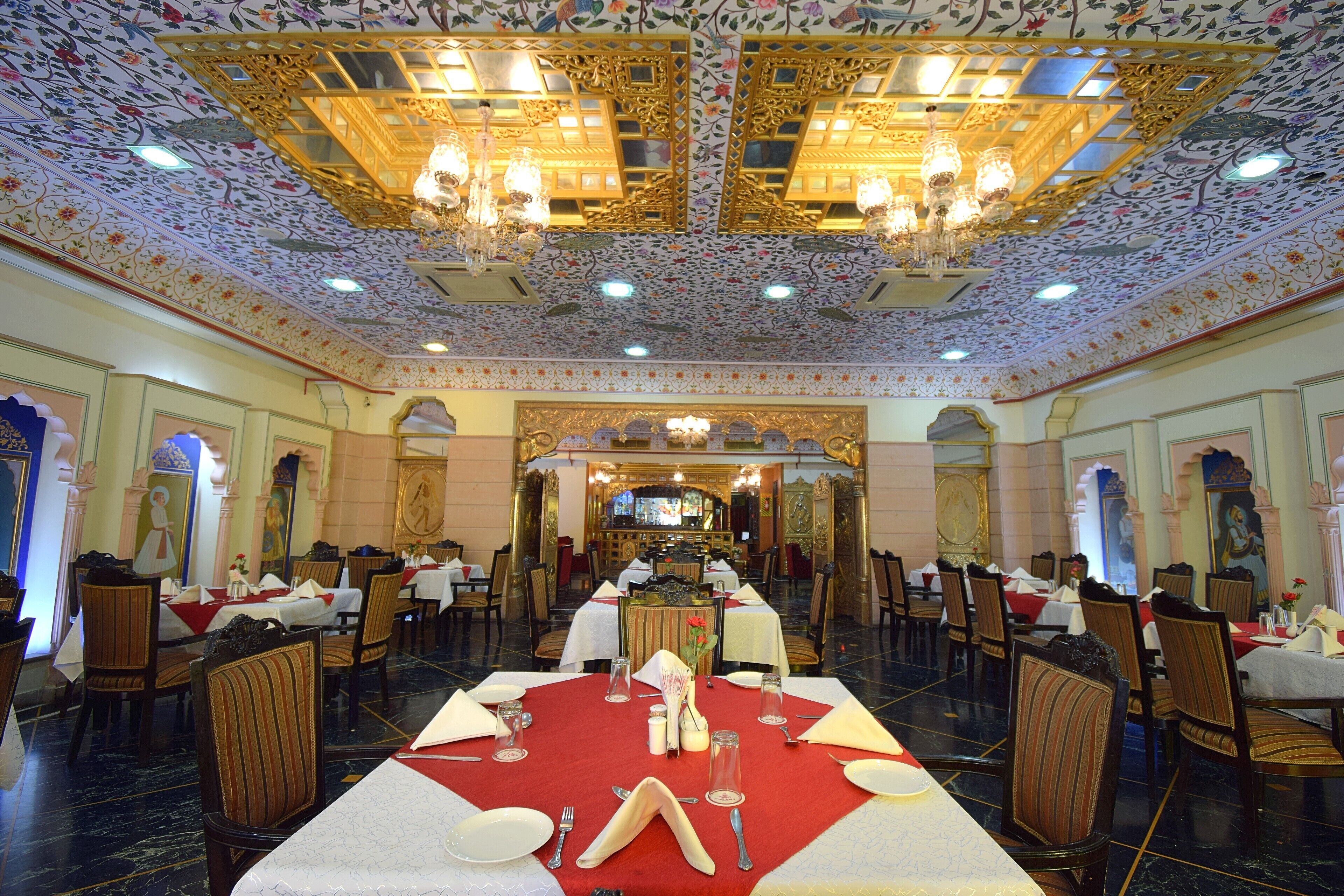 A dining area with tables set for four, ornate chandeliers, and a patterned ceiling.