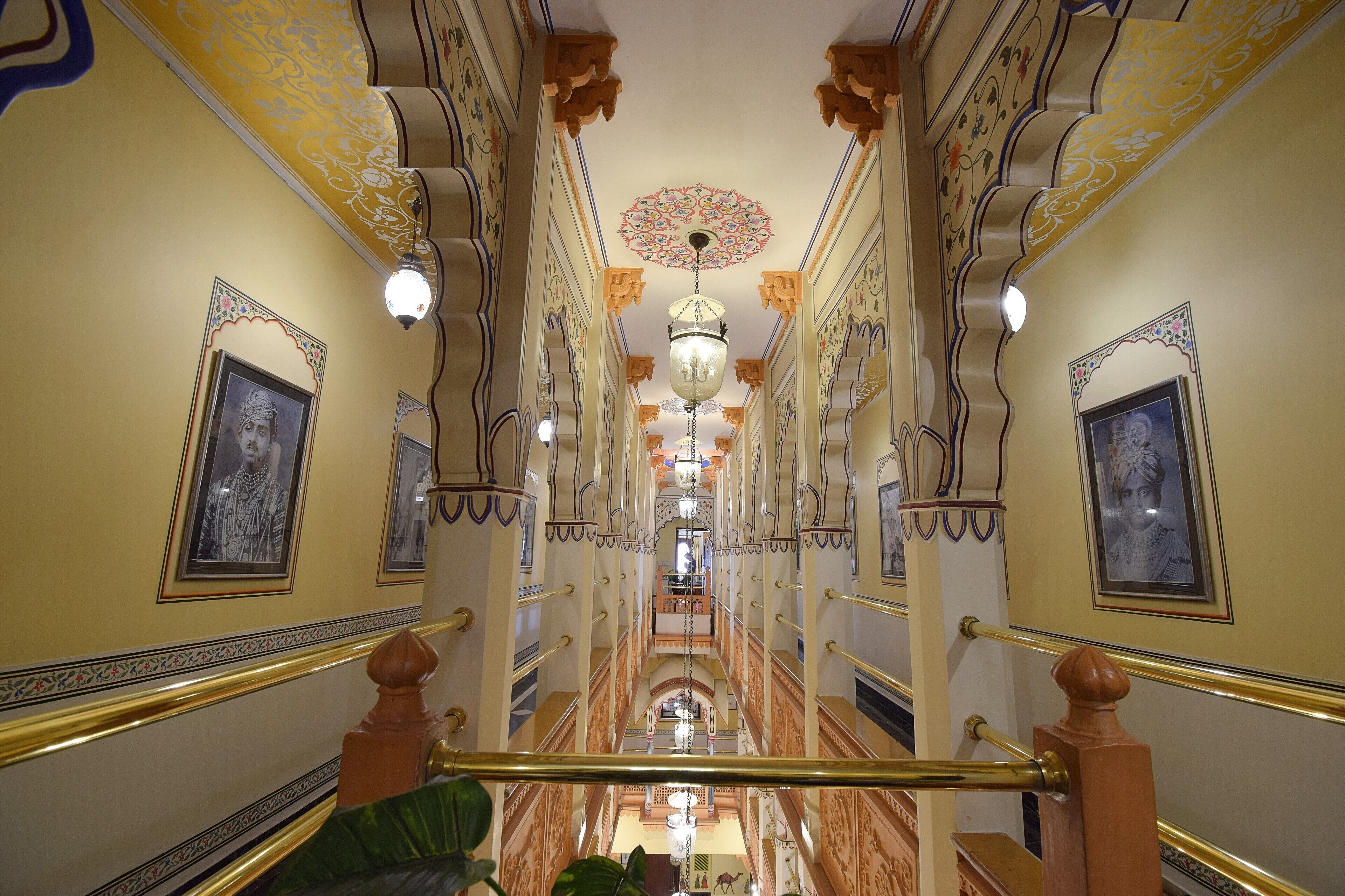 A grand, ornate hallway with a central chandelier, flanked by framed portraits and intricate wall designs.