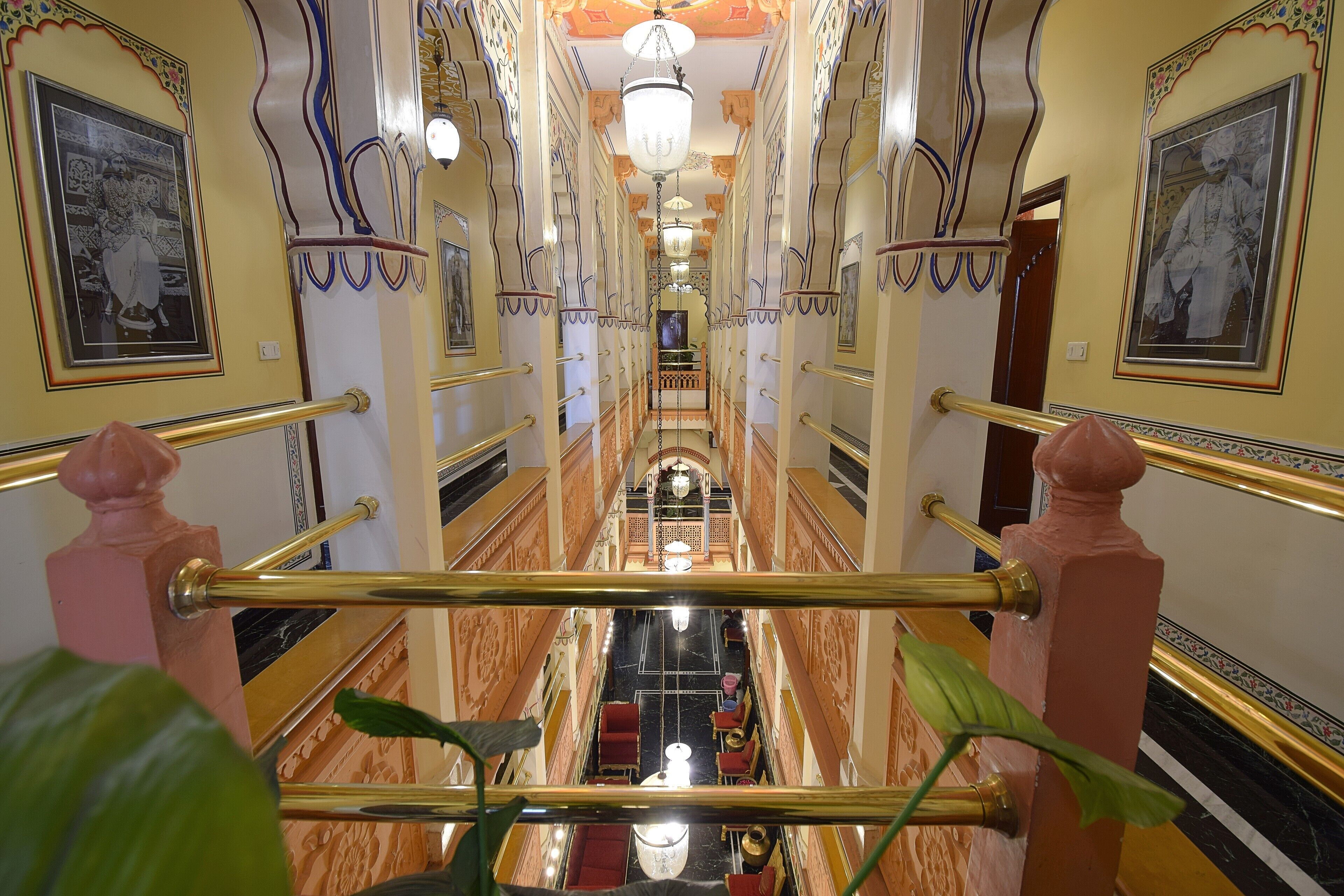 A grand hallway with ornate railings, chandeliers, and framed portraits on the walls.