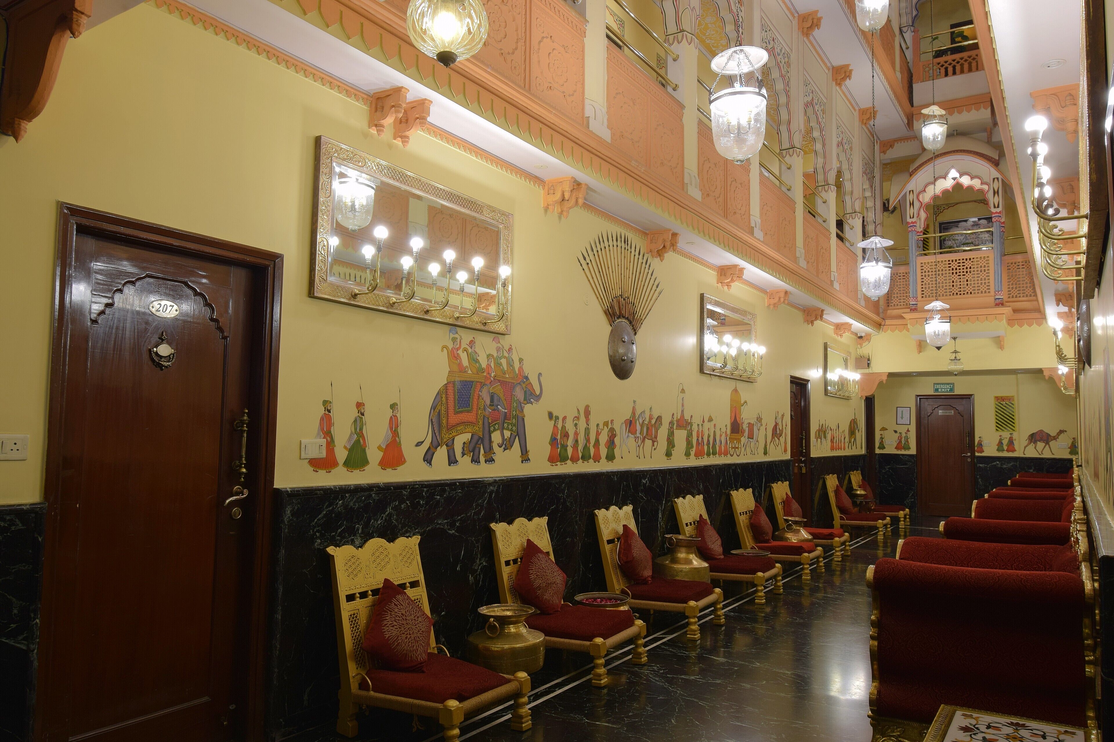Een hotelhal met rijen rode fluweelstoelen, een marmeren vloer en versierde wanddecoraties, waaronder een spiegel en kunstwerken met afbeeldingen van olifanten en mensen.
