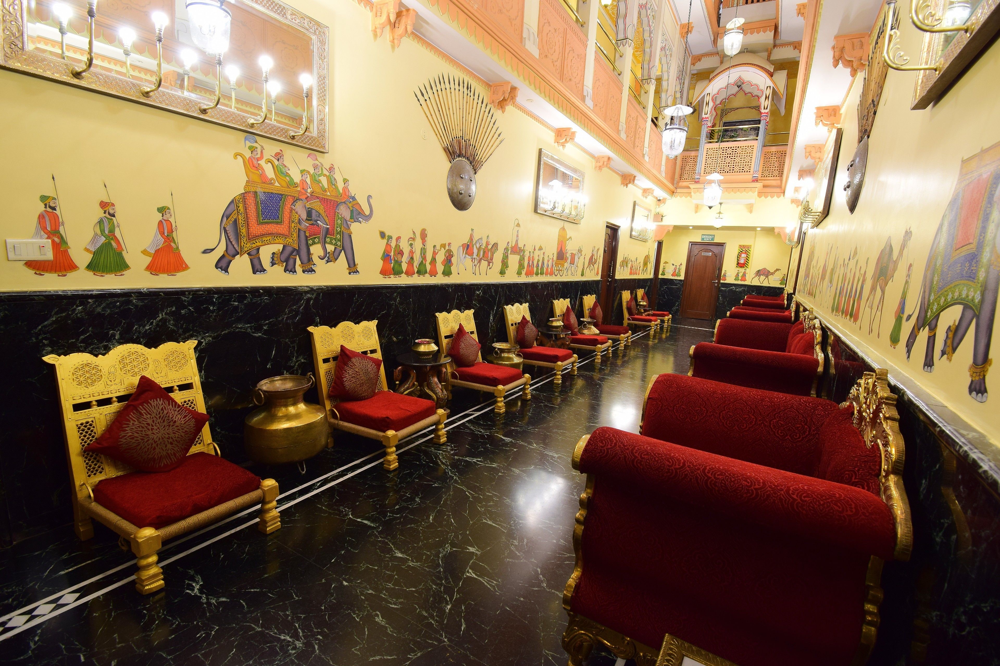 A hotel lobby with red upholstered chairs, a marble floor, and traditional Indian wall art.