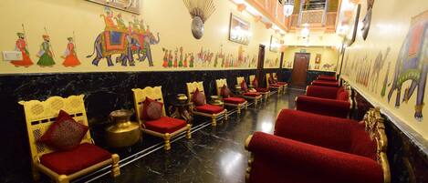 A hotel lobby with red upholstered chairs, a marble floor, and traditional Indian wall art.