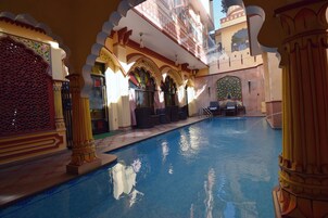 Piscina interna com detalhes arquitetônicos elaborados, incluindo arcos e colunas, e uma área de estar com cadeiras e uma mesa.
