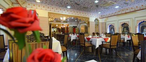 Un restaurante con techo ornamentado y candelabros, mesas dispuestas para comer y un jarrón con una rosa roja al frente.