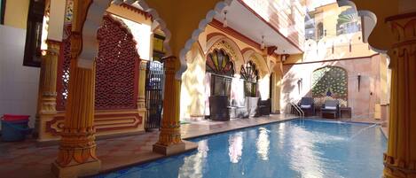 An indoor swimming pool with ornate arches and columns, a seating area with chairs and a table, and a decorative fountain.