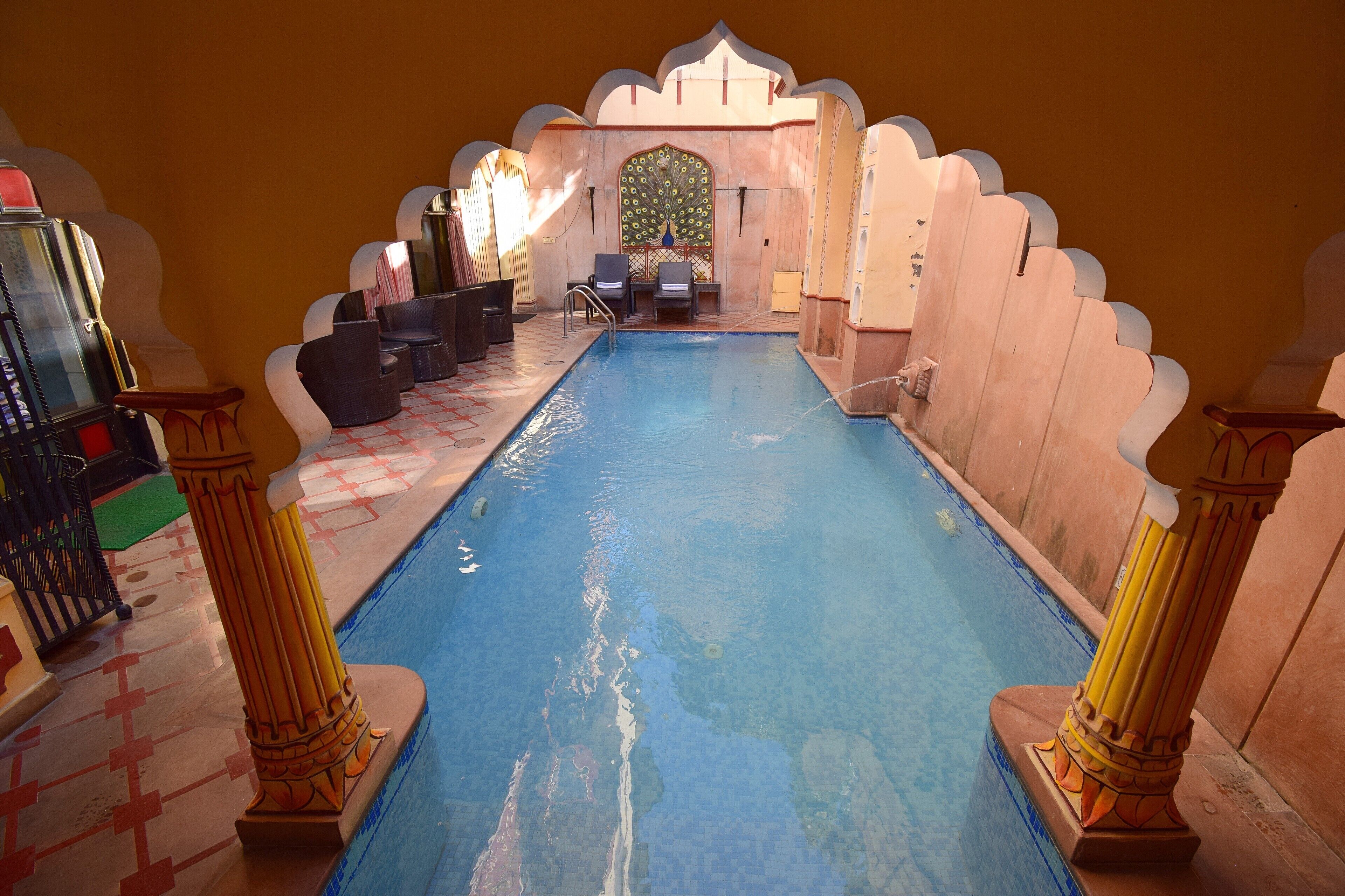 An indoor swimming pool with a peacock-themed wall, a seating area, and traditional architectural columns.