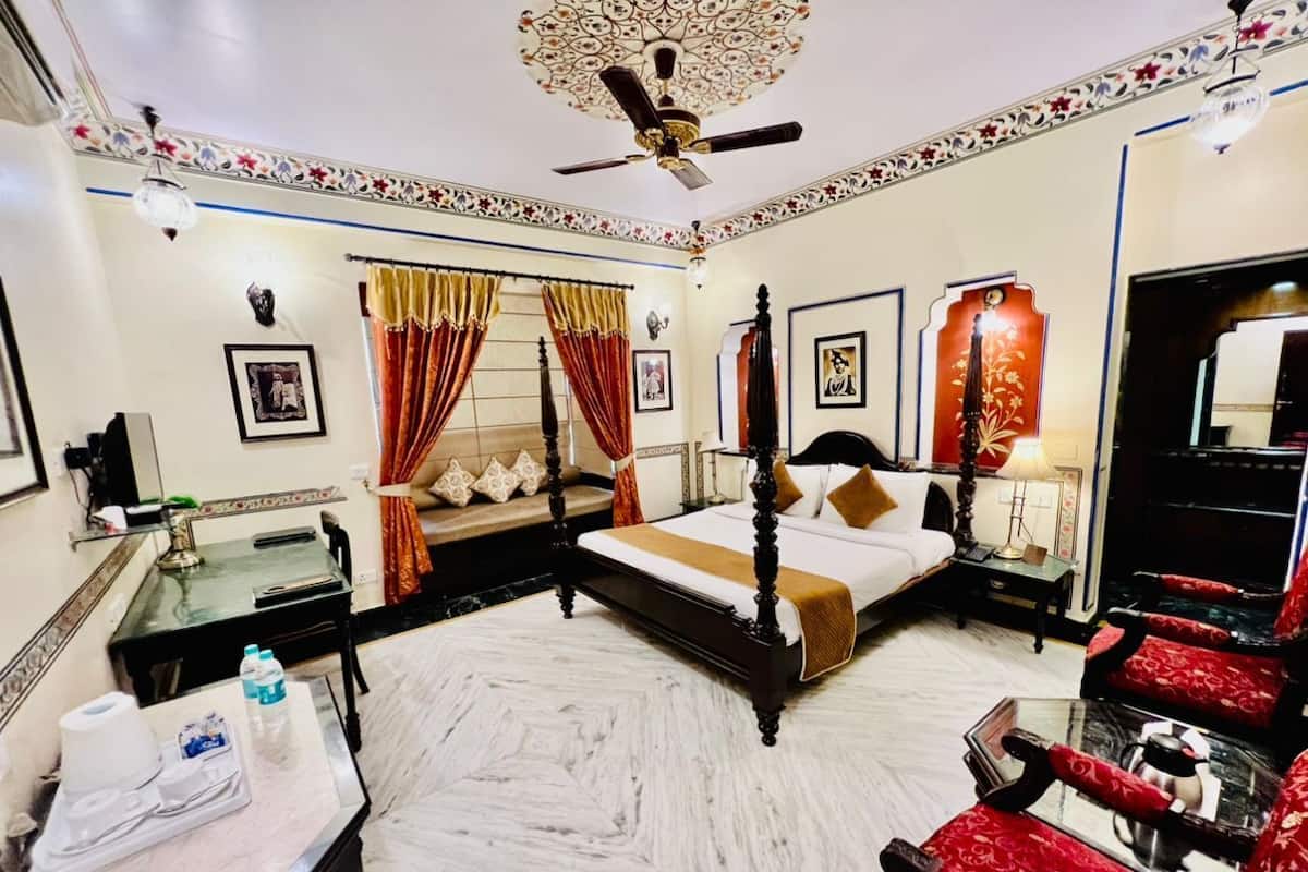 A hotel room with a four-poster bed, a sitting area with a red upholstered chair, a small table with a lamp, and a ceiling fan.