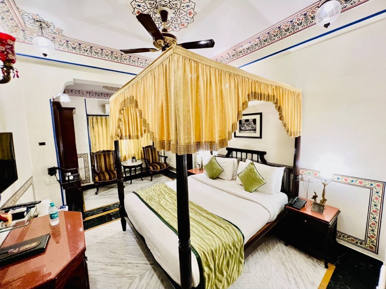 A four-poster canopy bed with a green throw, a nightstand with a lamp, and a chair in a hotel room.