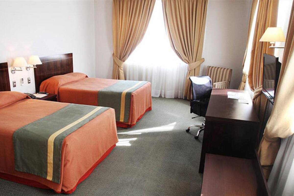 A hotel room with two beds, a desk, a chair, a lamp, and a window with curtains.