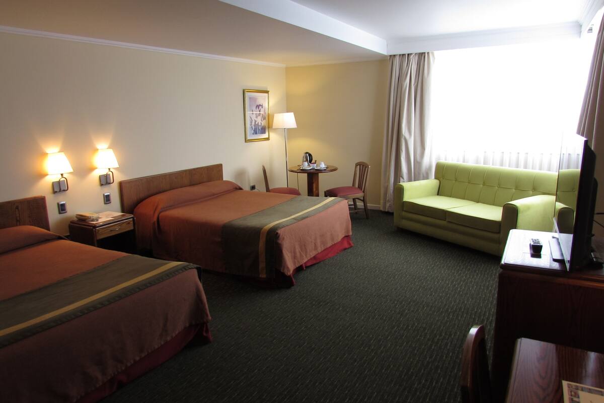 A hotel room with two beds, a sofa, a desk with a computer, a lamp, and a window with curtains.