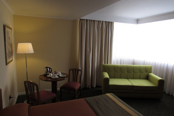 A hotel room with a bed, a green sofa, a small table with a lamp, and a window with curtains.