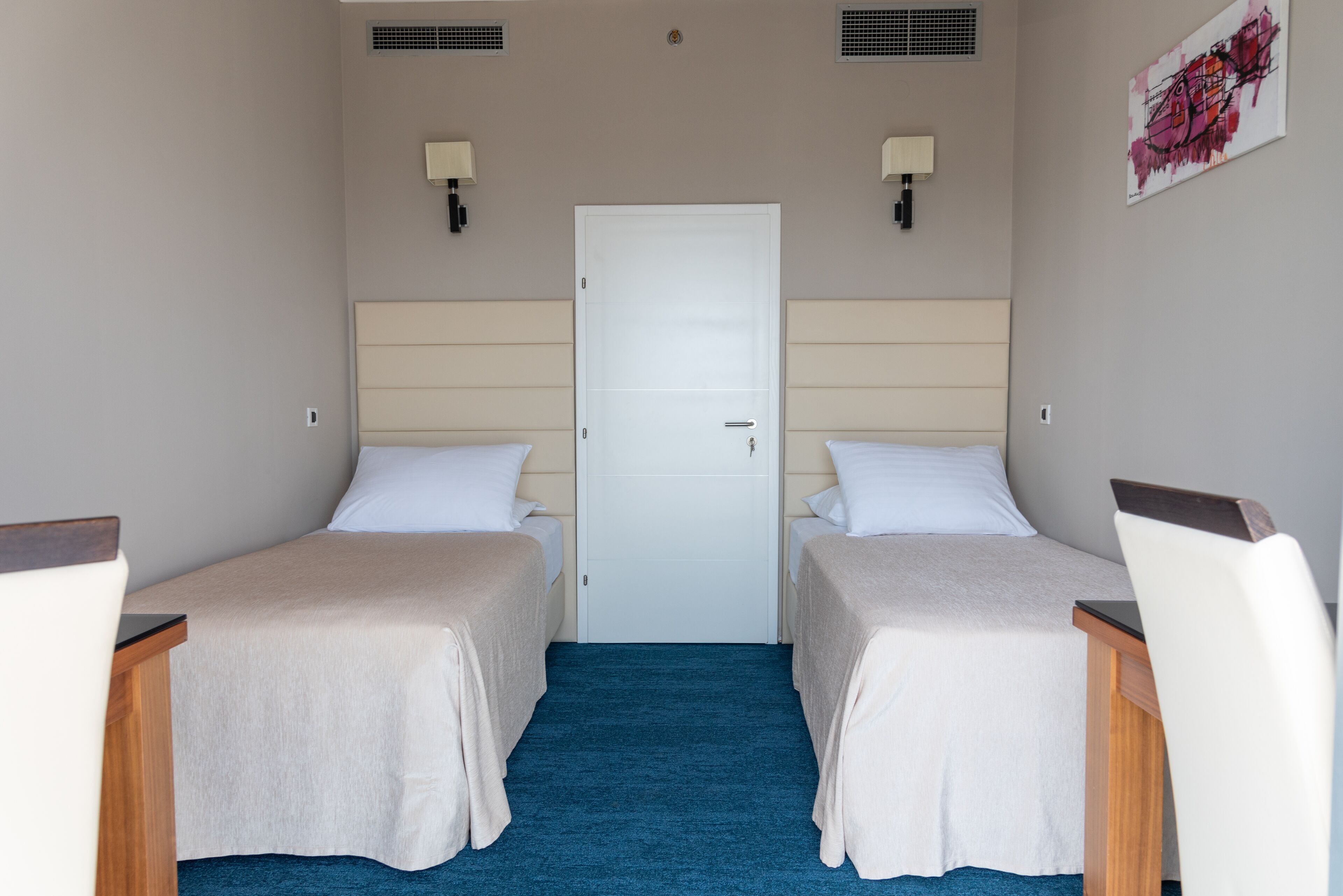 A hotel room with two single beds, a wooden desk, a chair, a blue carpet, a white door, and two wall-mounted lamps.