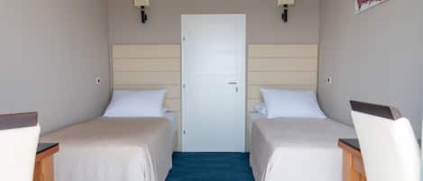 A hotel room with two single beds, a wooden desk, a chair, a blue carpet, a white door, and two wall-mounted lamps.