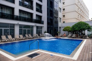 Piscina com espreguiçadeiras e cascata, emoldurada por edifícios modernos.