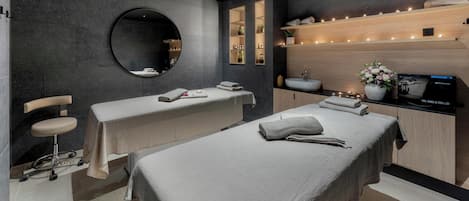 A modern spa room with two treatment tables, a massage chair, and a wooden vanity with a sink and towel warmer.