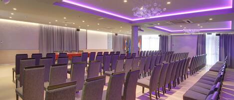 A conference room with rows of chairs, a stage, and a large screen.