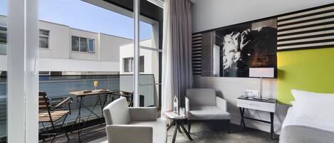 Habitación de hotel moderna con un ventanal grande, un rincón de comedor con mesa y sillas, una cama, una mesita de noche con lámpara y un cuadro en la pared.