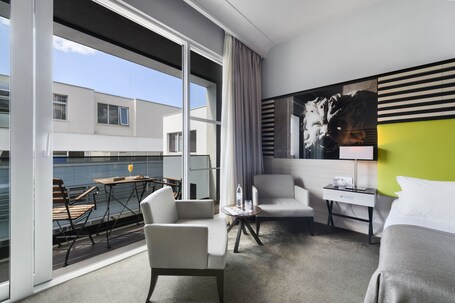 A modern hotel room with a large window, a dining area with a table and chairs, a bed, a small table with a lamp, and a painting on the wall.
