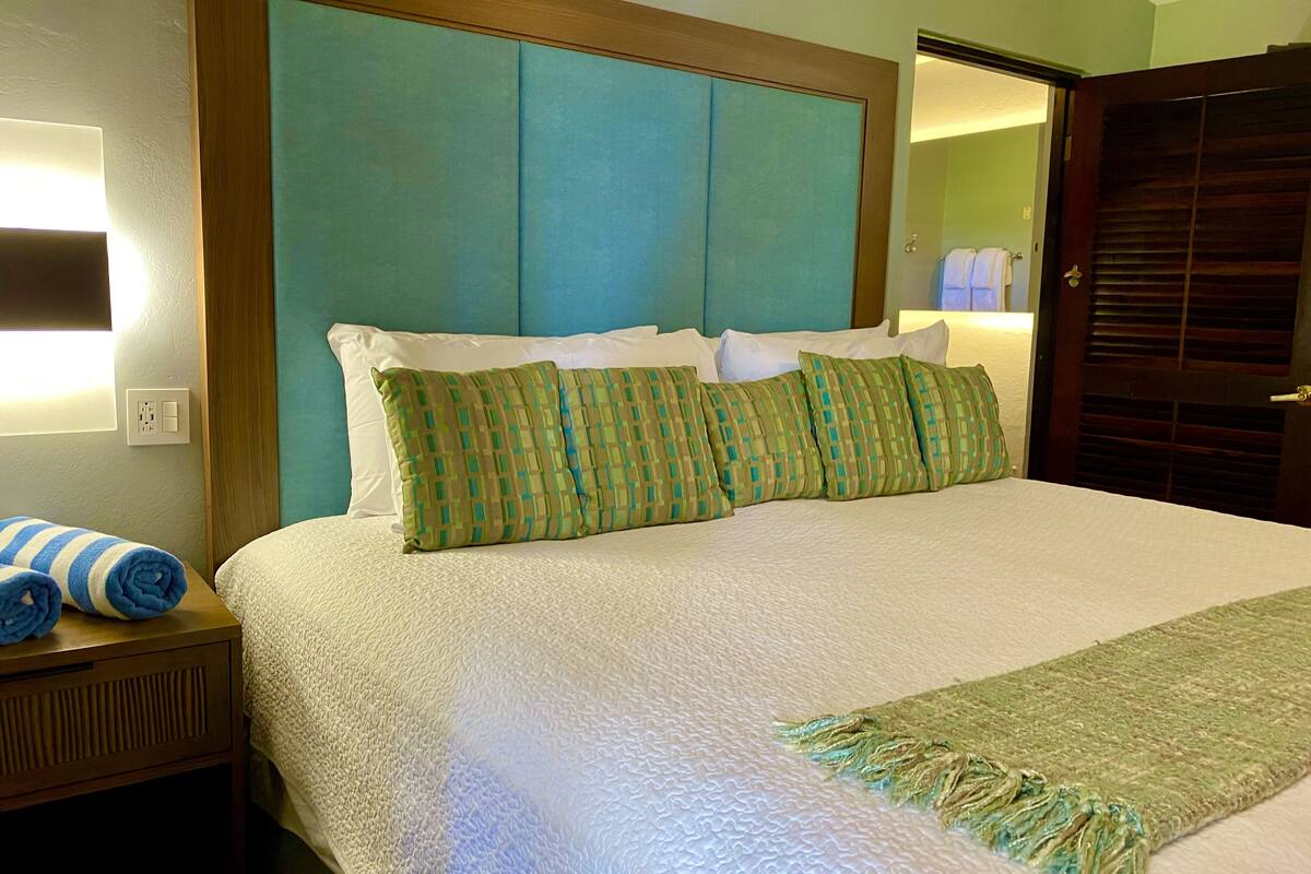 Een netjes opgemaakt bed met een witte deken en groene kussens met een patroon, een houten nachtkastje met een opgerolde handdoek en een groene plaid over het bed.