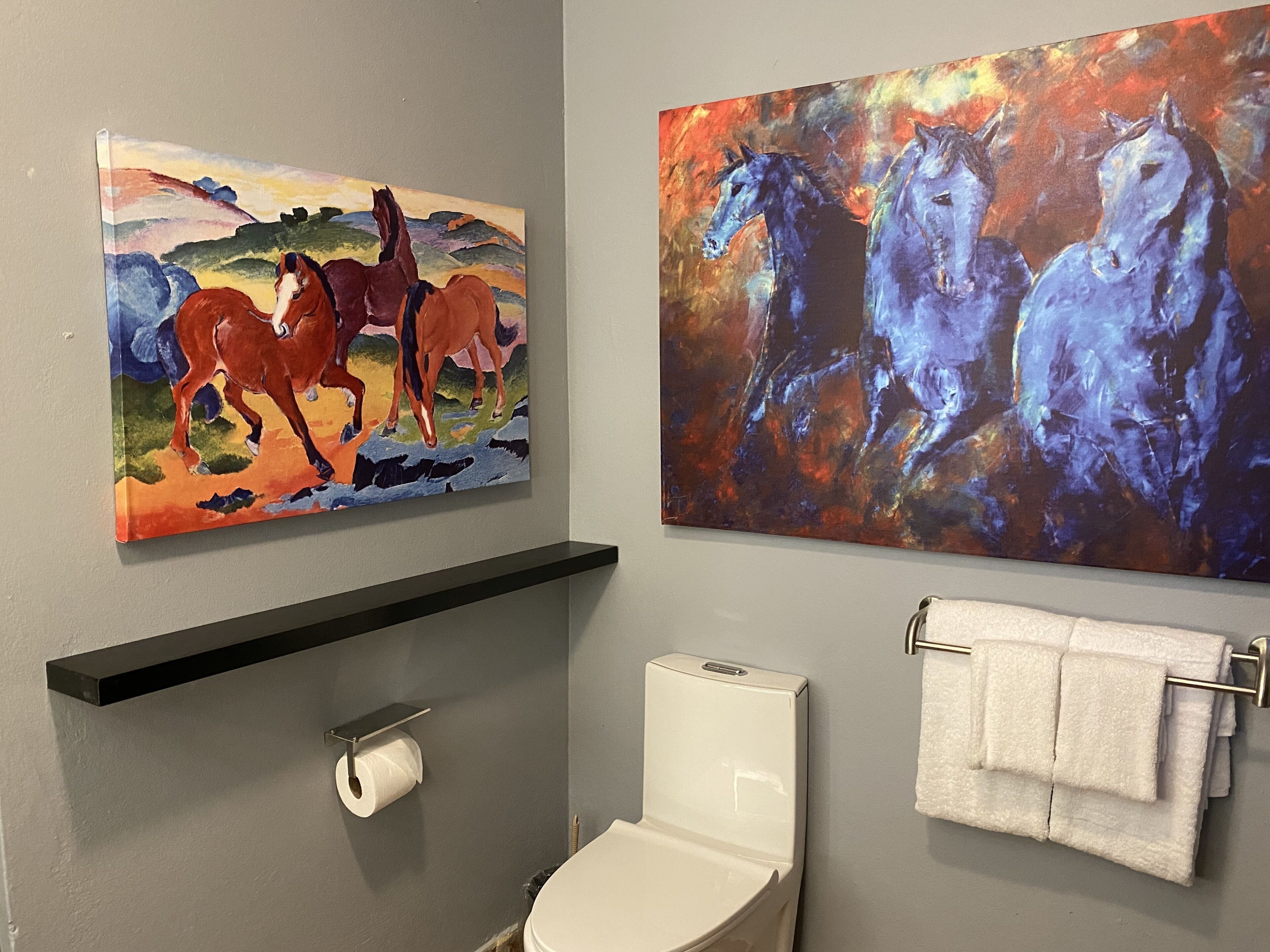 Une salle de bain équipée de toilettes, d’un porte-serviettes et de deux tableaux représentant des chevaux.