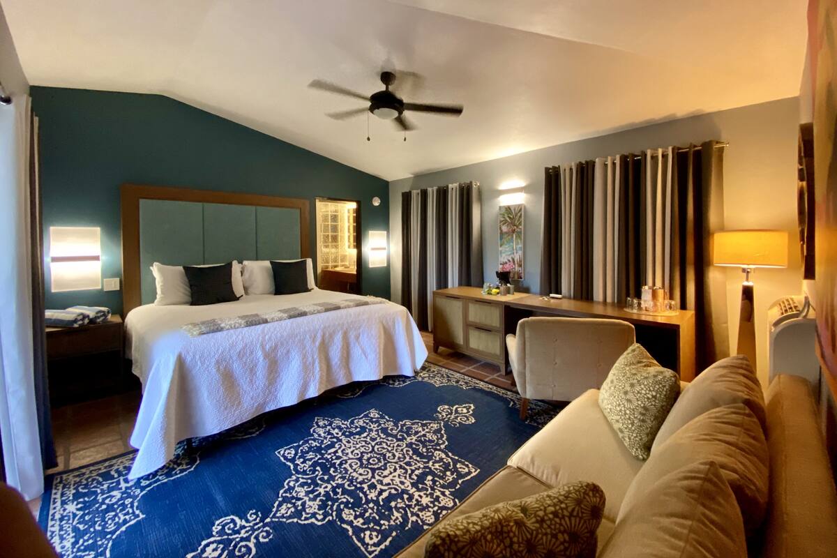 Hotelkamer met een bed, bureau, stoel, plafondventilator en een groot raam met gordijnen.