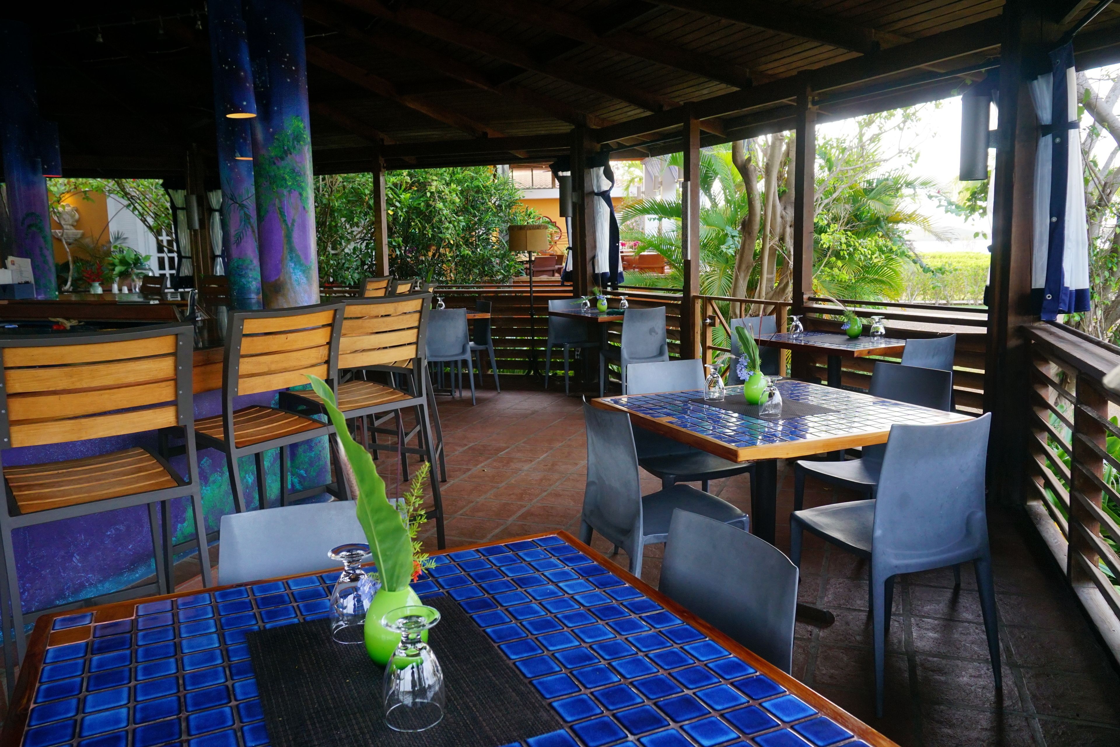 Een restaurant buiten met houten tafels en stoelen, een overdekt terras en uitzicht op groen.