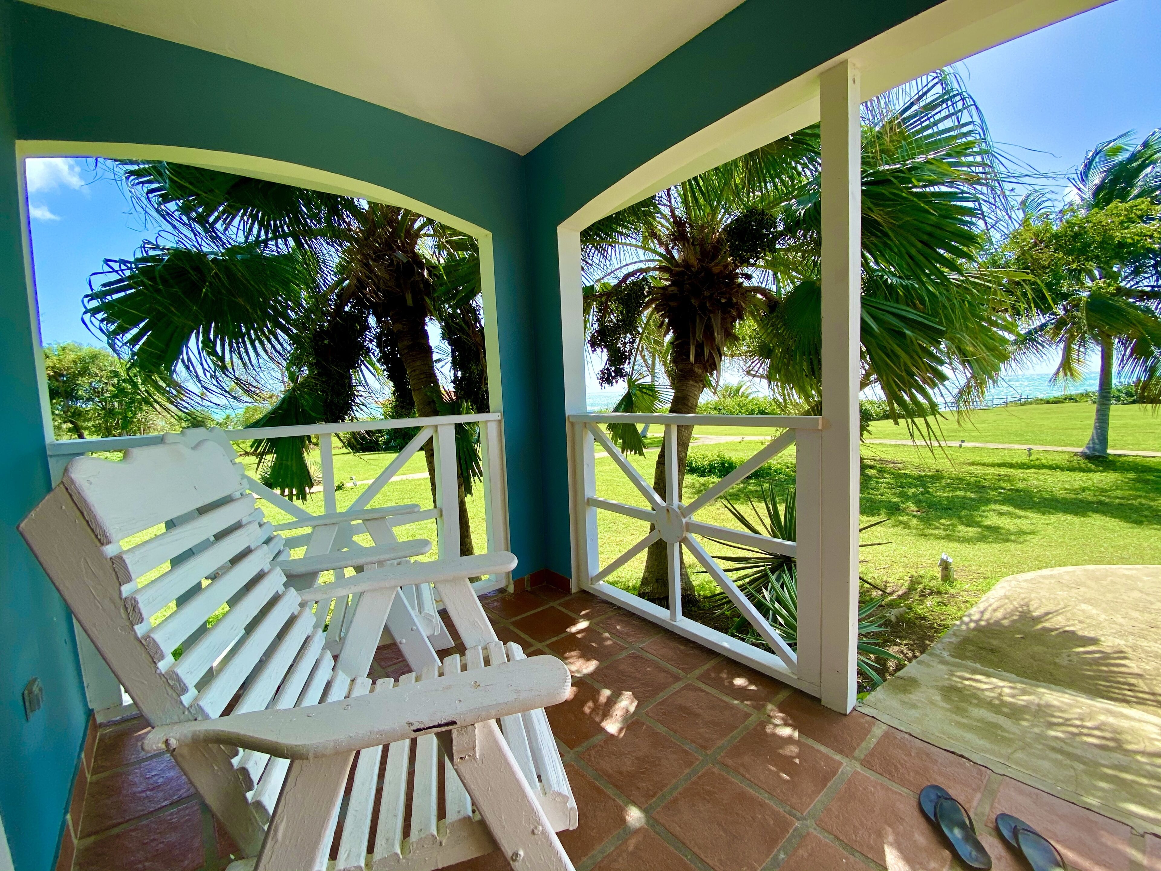 A covered outdoor seating area with a bench, a view of a lawn with palm trees, and a clear sky.