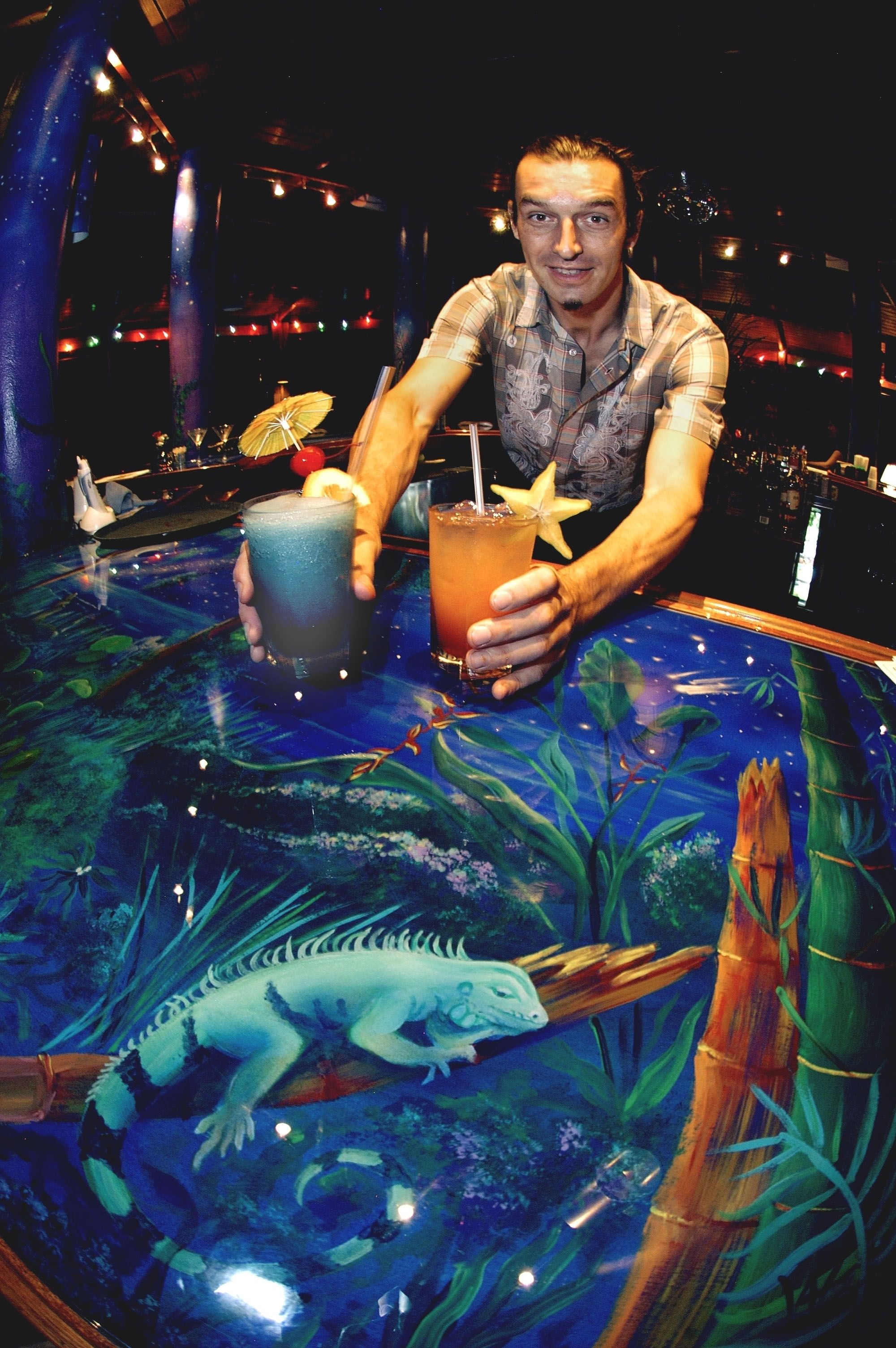 Pessoa segurando duas bebidas em um bar com mesa de vidro com desenho de tartaruga marinha.