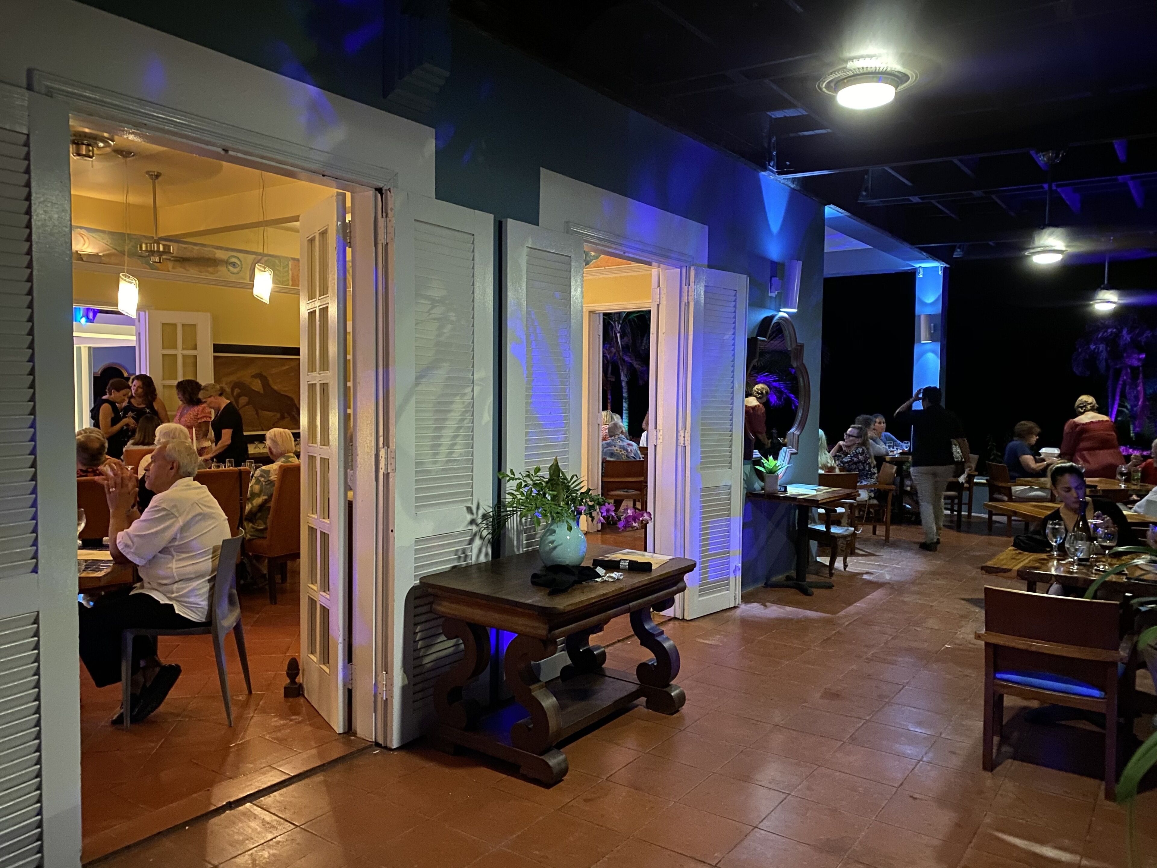 An indoor dining area with open doors leading to a kitchen, wooden tables, and chairs.