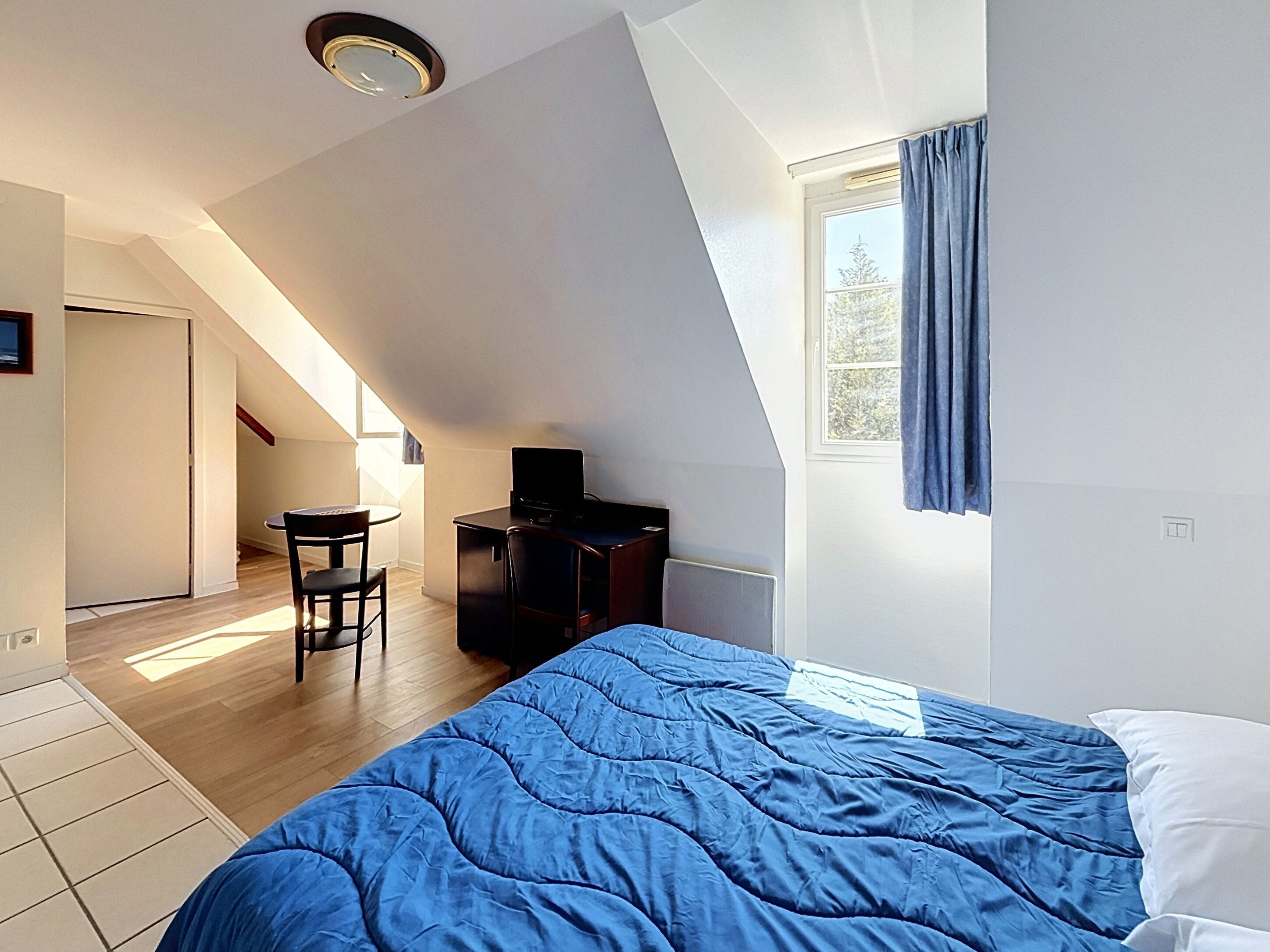 A bedroom with a sloped ceiling, a bed with a blue duvet, a desk with a computer, and a window with curtains.