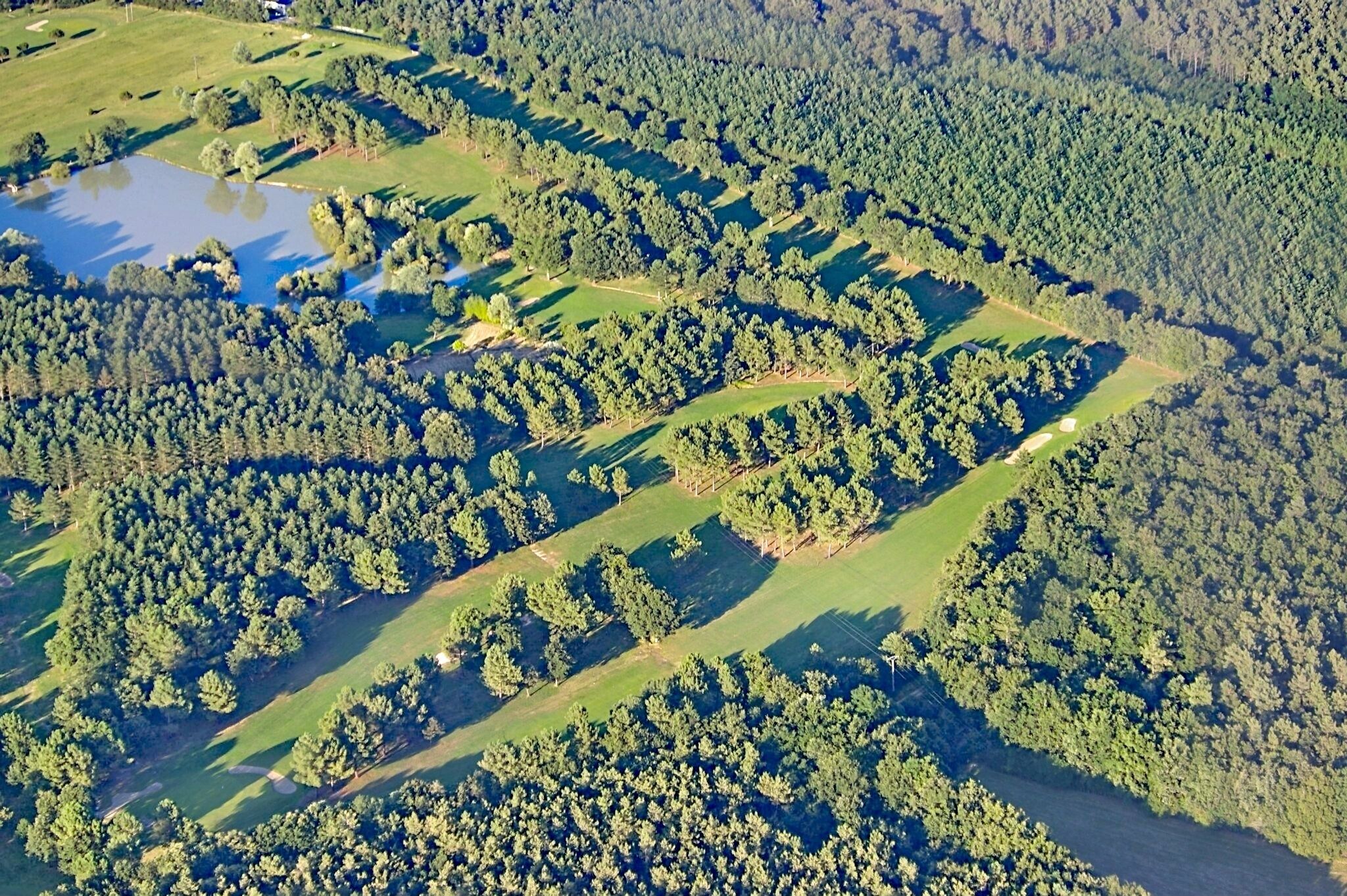 An aerial view of a golf course with multiple greens, a pond, and dense forested areas.