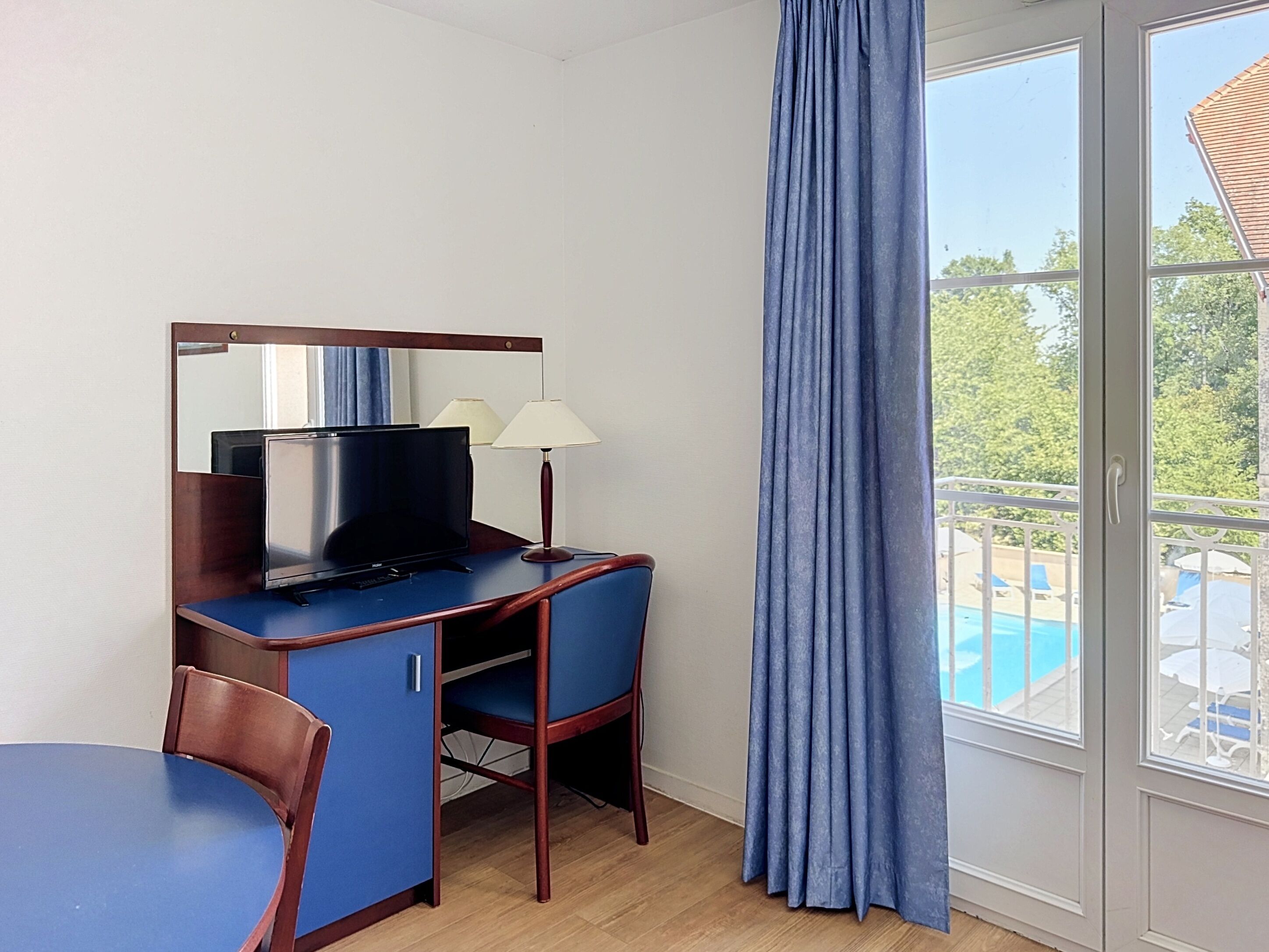 A hotel room with a desk, television, two lamps, a chair, and a view of a pool.