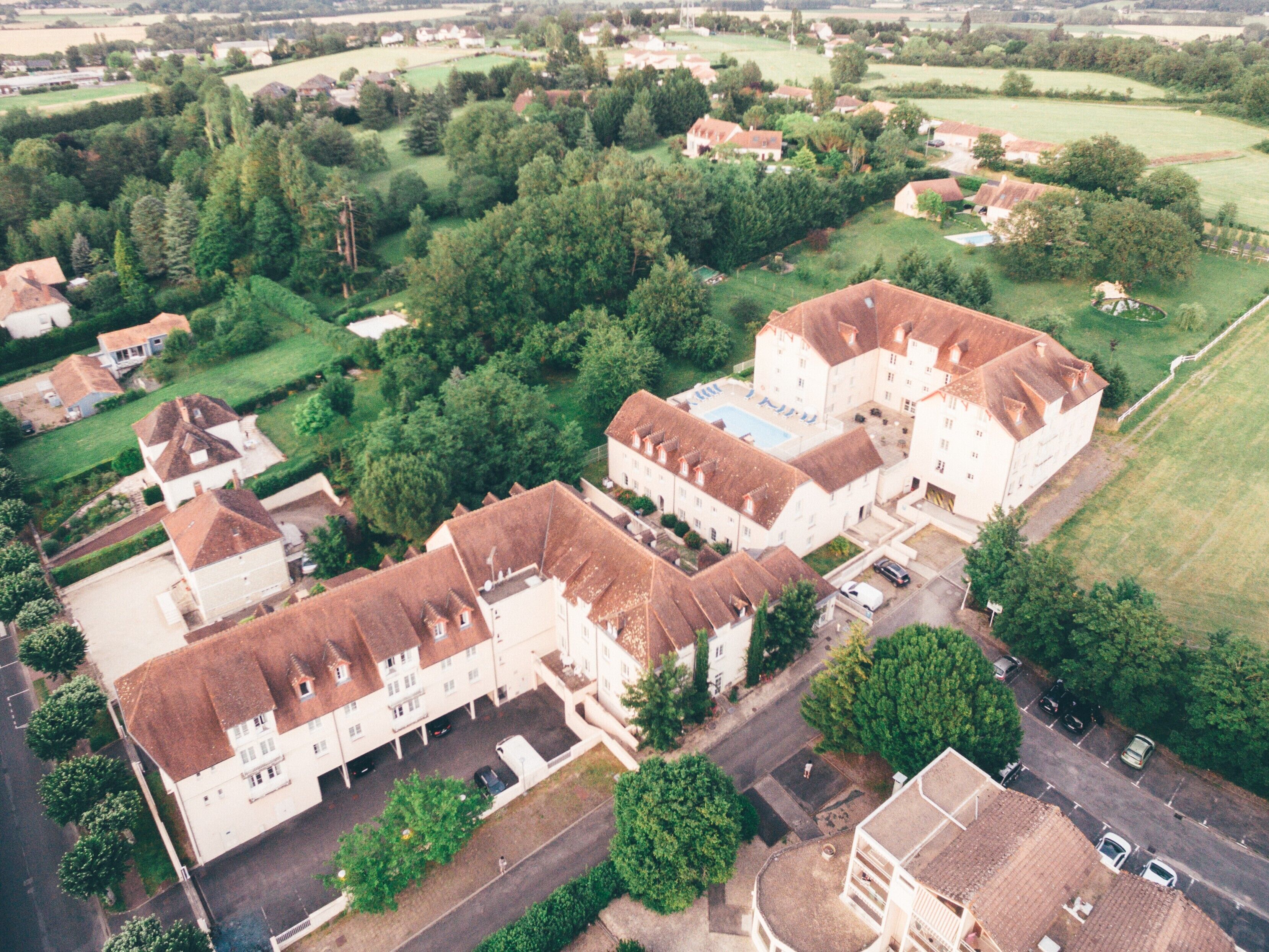 An aerial view of a residential area with multiple houses, a large building with a swimming pool, and surrounding greenery.