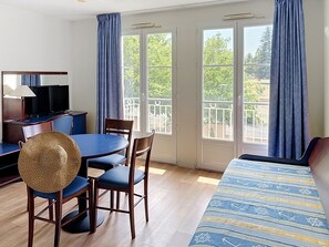 A hotel room with a bed, a desk with a TV, a dining table with chairs, and large windows with curtains. - Terres de France - Appart'Hôtel La Roche Posay (La Roche-Posay)
