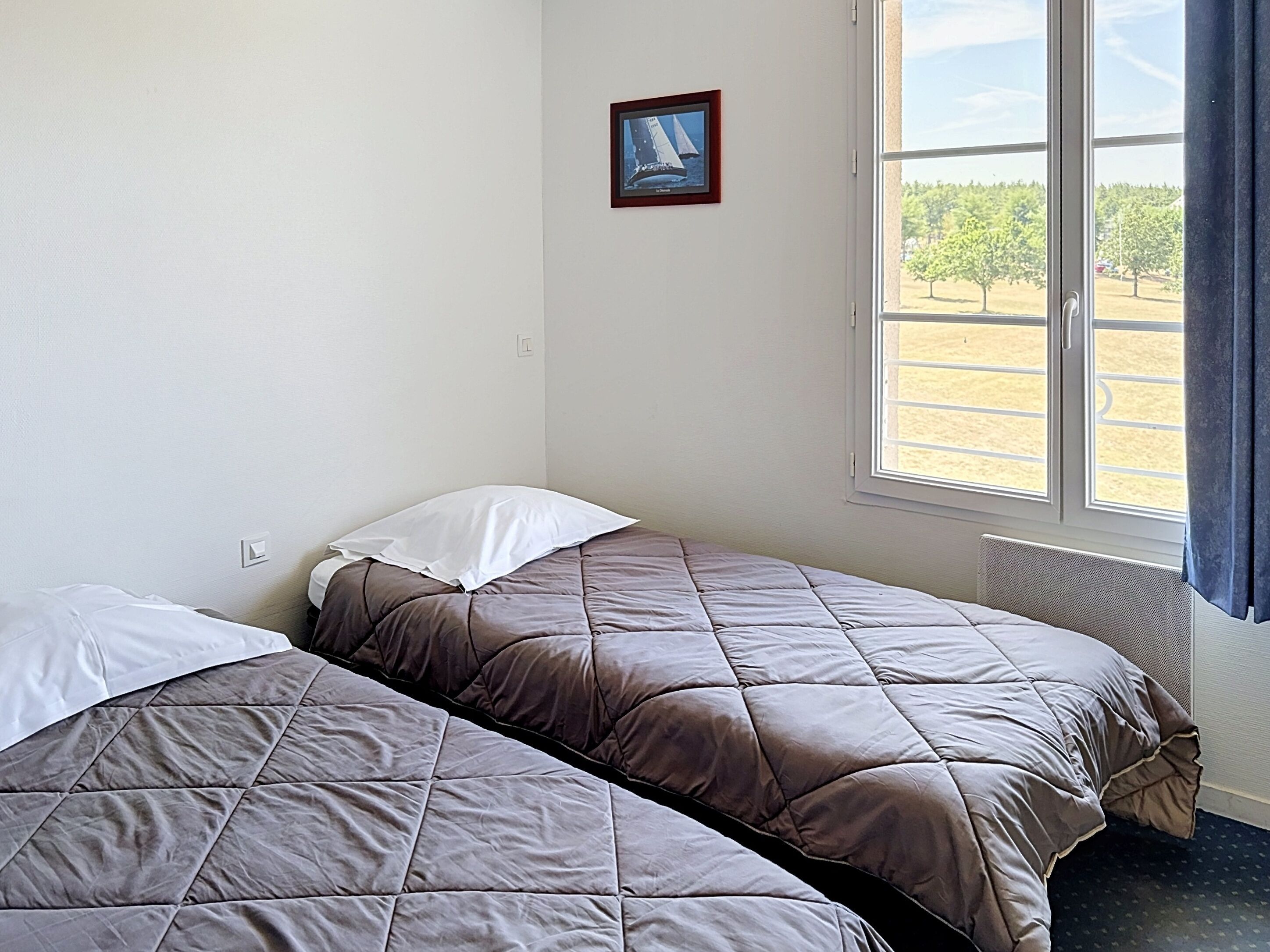 A room with two beds, a window with a view of a field, and a framed picture on the wall.