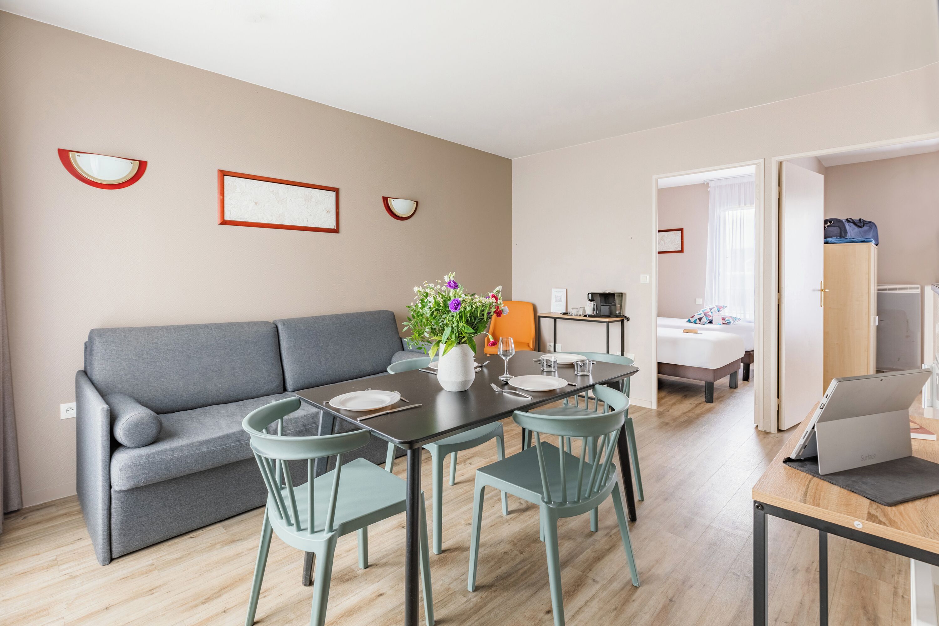 Ein modernes Wohnzimmer mit grauem Sofa, einem Esstisch mit Stühlen, einer kleinen Küchenzeile und einem durchgehenden Türrahmen einsehbaren Schlafzimmer.