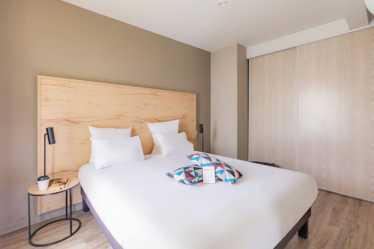 A modern hotel room with a large bed, a wooden headboard, a small bedside table with a lamp, and a wardrobe.