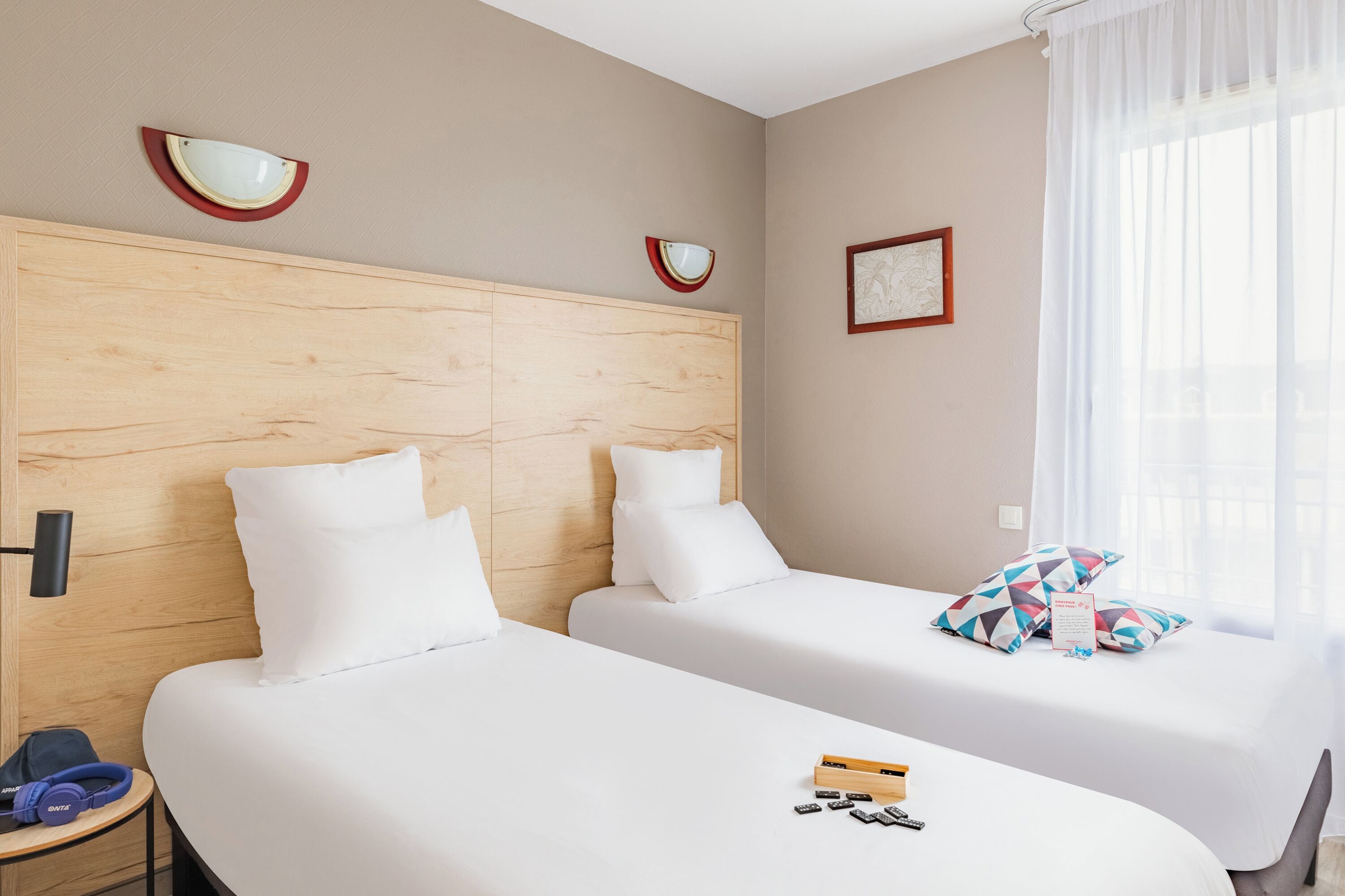 A hotel room with two beds, a wooden headboard, a small bedside table, a lamp, and a window with sheer curtains.