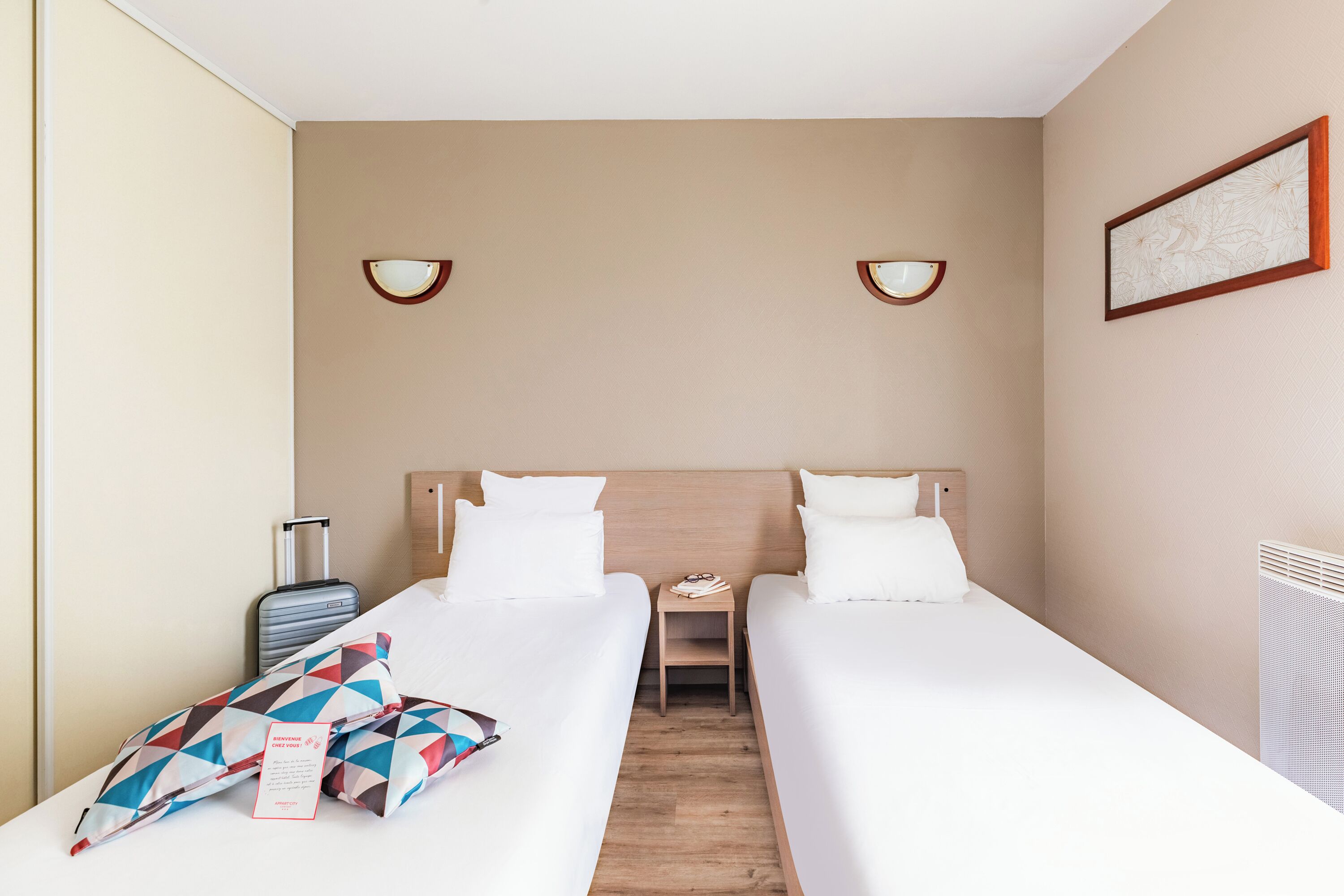 Habitación de hotel con dos camas individuales, una mesita de noche pequeña, una lámpara, una maleta y un cuadro en la pared.