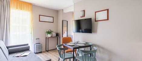 Un salon moderne avec un canapé, une table à manger et des chaises, un téléviseur fixé au mur et une fenêtre avec des rideaux.