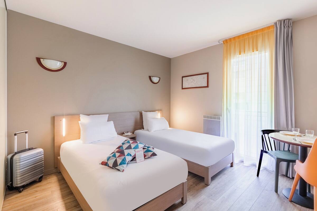 A hotel room with two beds, a desk with a chair, a suitcase, and a window with curtains.
