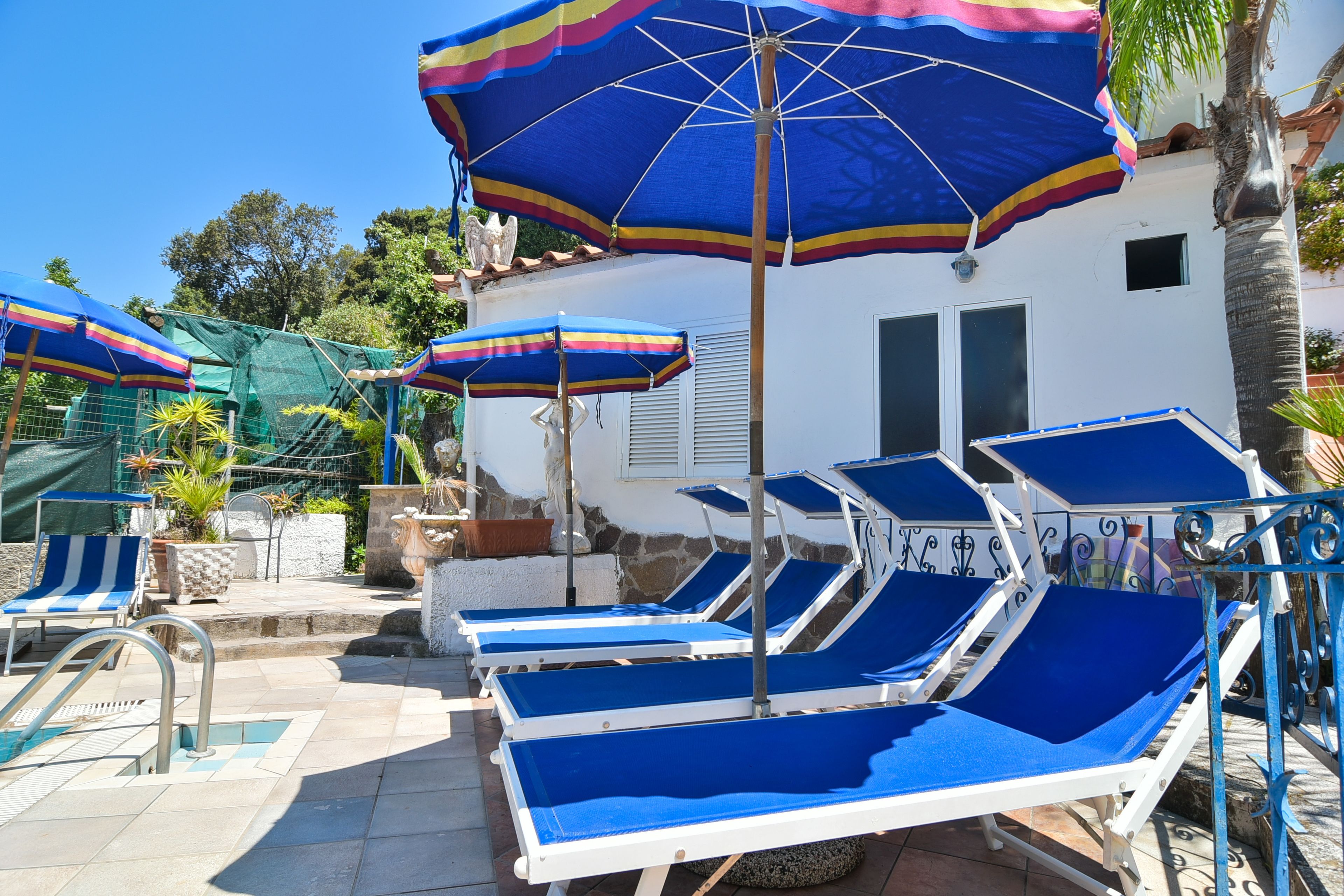 Un espace aménagé au bord de la piscine, avec des chaises longues bleues et des parasols colorés, un mur en pierre et un bâtiment blanc doté de volets.