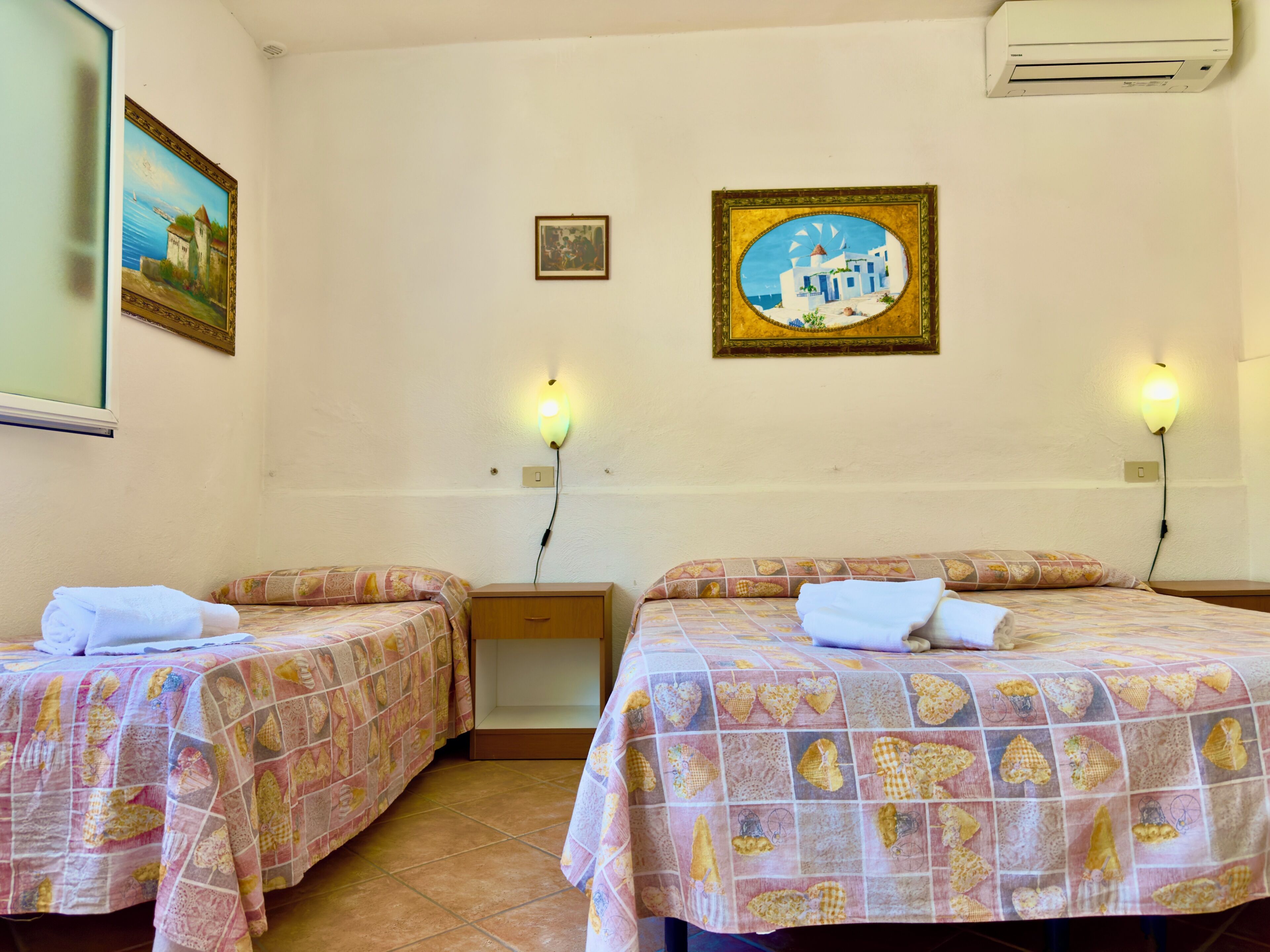 A hotel room with two beds, each with a patterned bedspread, a small wooden nightstand, a wall-mounted air conditioner, and two wall-mounted light fixtures.