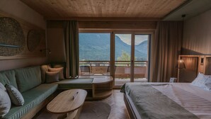 A modern hotel room with a wooden ceiling, a large bed, a sofa, a small table, and a balcony with a mountain view. - Die Wasnerin G'sund & Natur Hotel (Bad Aussee)