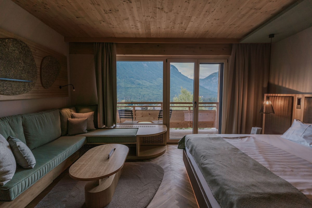 Habitación de hotel moderna con techo de madera, una cama grande, un sofá, una mesita y un balcón con vistas a la montaña.