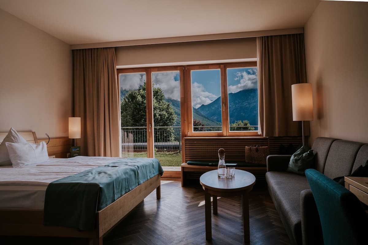 Habitación de hotel con cama, sofá, una mesita y vistas a montañas a través de amplios ventanales.