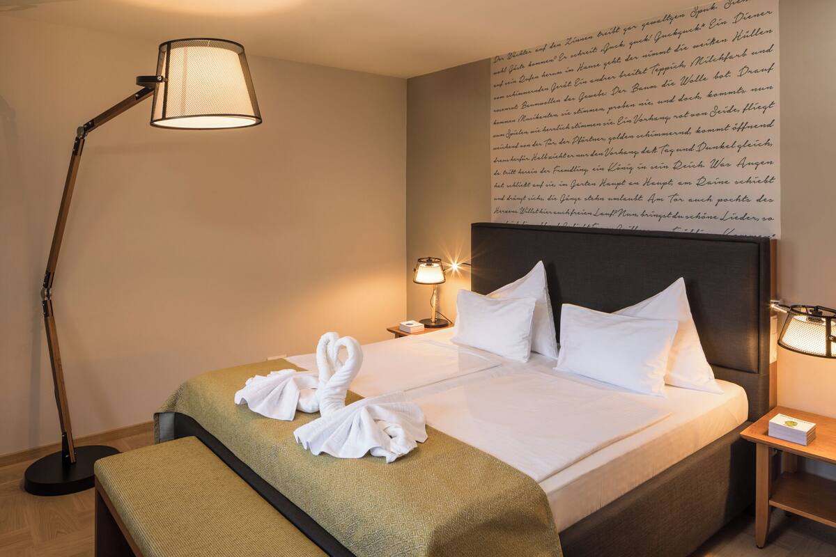 Una habitación de hotel con una cama bien tendida, una mesita de noche con una lámpara y una pared con un gran texto.